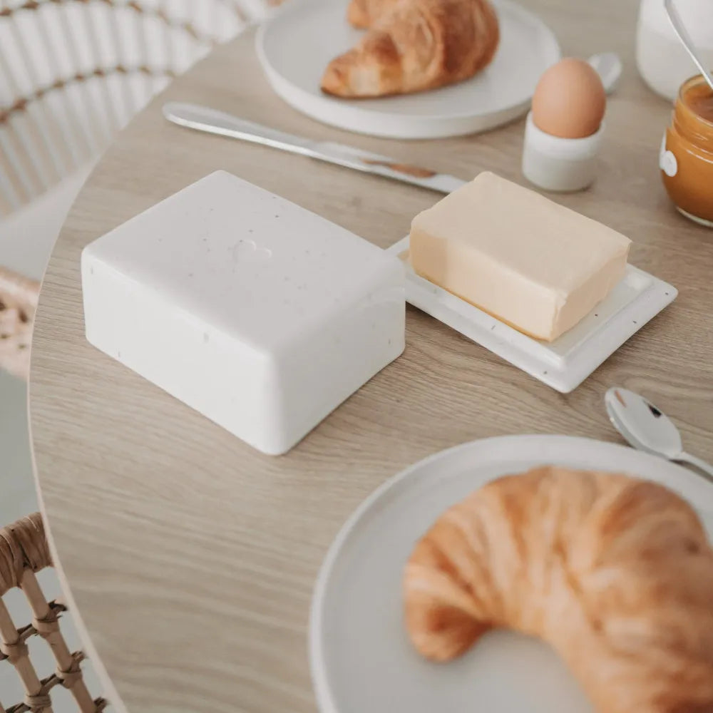 Offene helle Butterdose zwischen Croissants auf Tellern, Ei und Aufstrich auf einem Holztisch