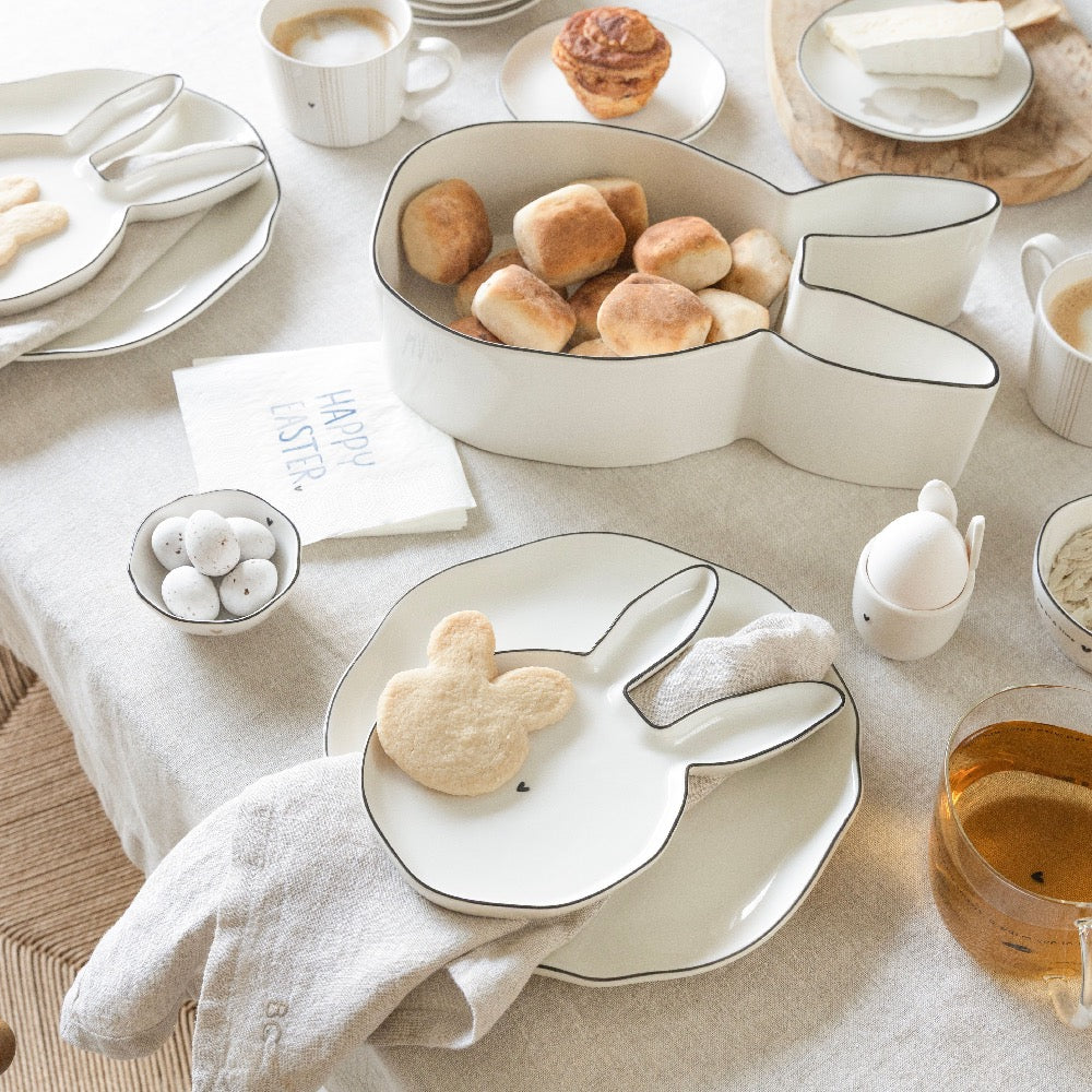 Ein gedeckter Tisch mit Tellern in Hasenform, einer großen Hasenschale mit Brötchen, Kaffeetassen, einer Serviette mit der Aufschrift "Frohe Ostern" und verschiedenen Frühstücksgerichten auf einer neutralen Tischdecke.