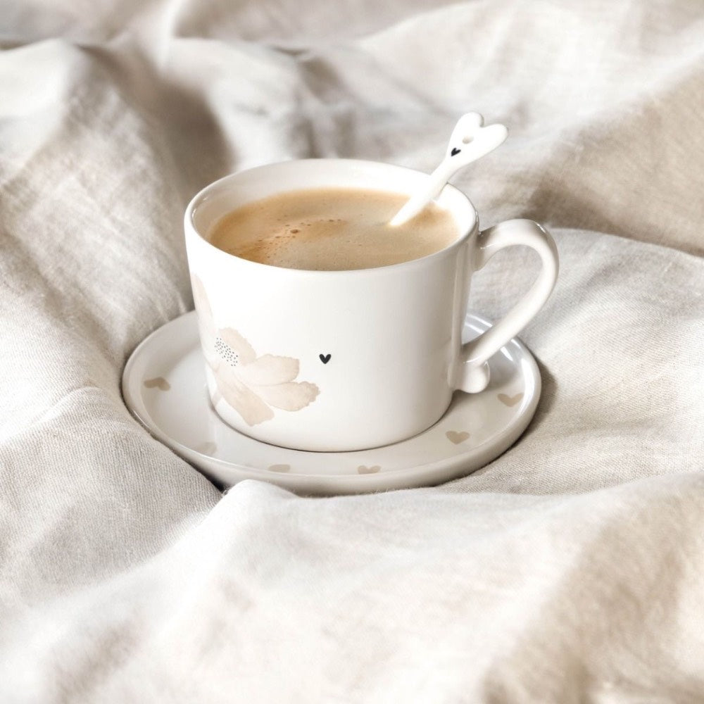 Eine weiße Kaffeetasse mit Herzmuster und ein herzförmiger Löffel sitzen auf einer passenden Untertasse auf einer beigen Stoffoberfläche.