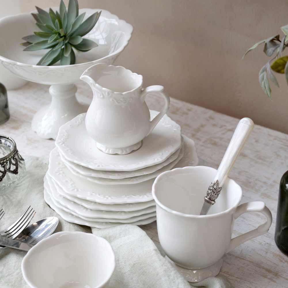 Weißes Provence-Geschirr mit Tellerstapel, Milchkännchen und Tasse auf einem hellen Tisch