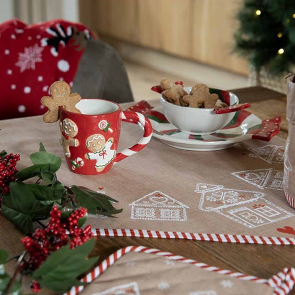Rote Tasse mit Lebkuchenmotiv auf einem Tisch mit gemusterten Textilien und Gebäck