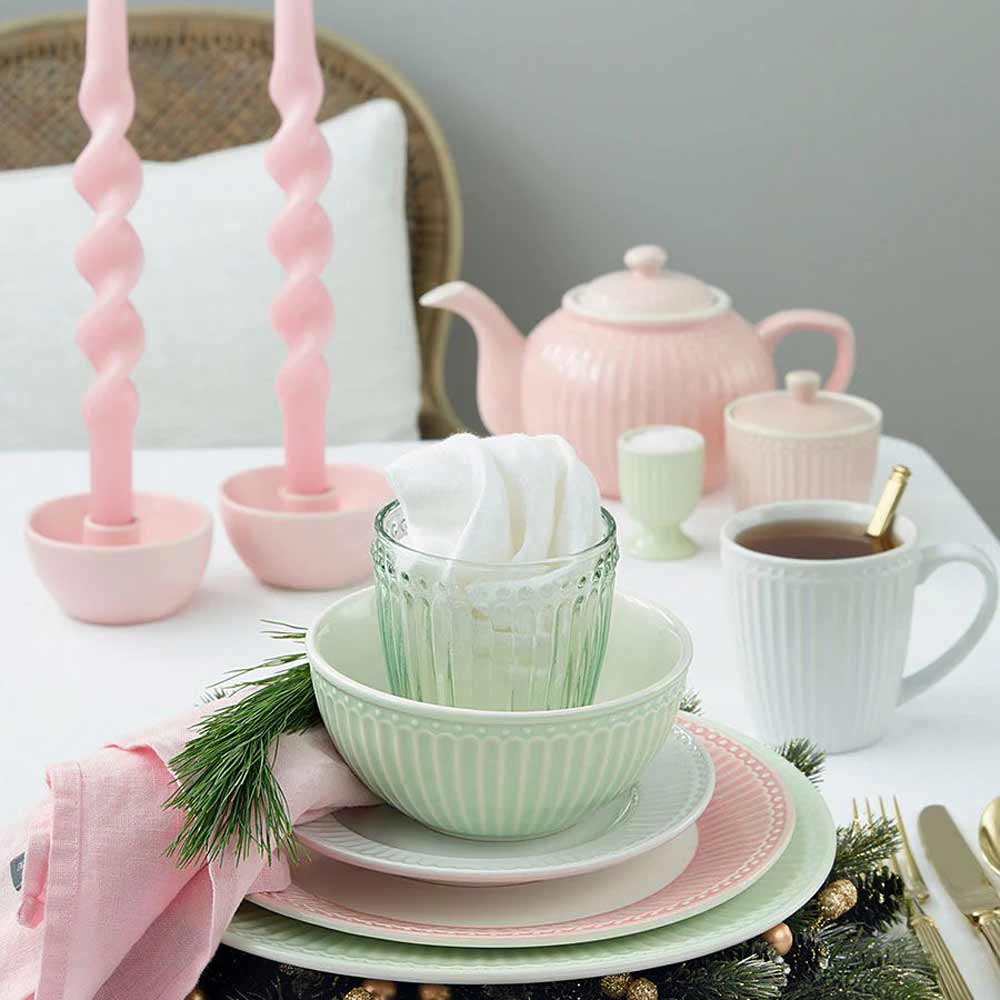 Auf einem weißen Tisch mit Tannenzweigen und einer rosa Serviette sind pastellfarbenes Geschirr, eine Teekanne, spiralförmige rosa Kerzen und eine Tasse Tee angeordnet, wodurch eine festliche und elegante Tischdekoration entsteht.