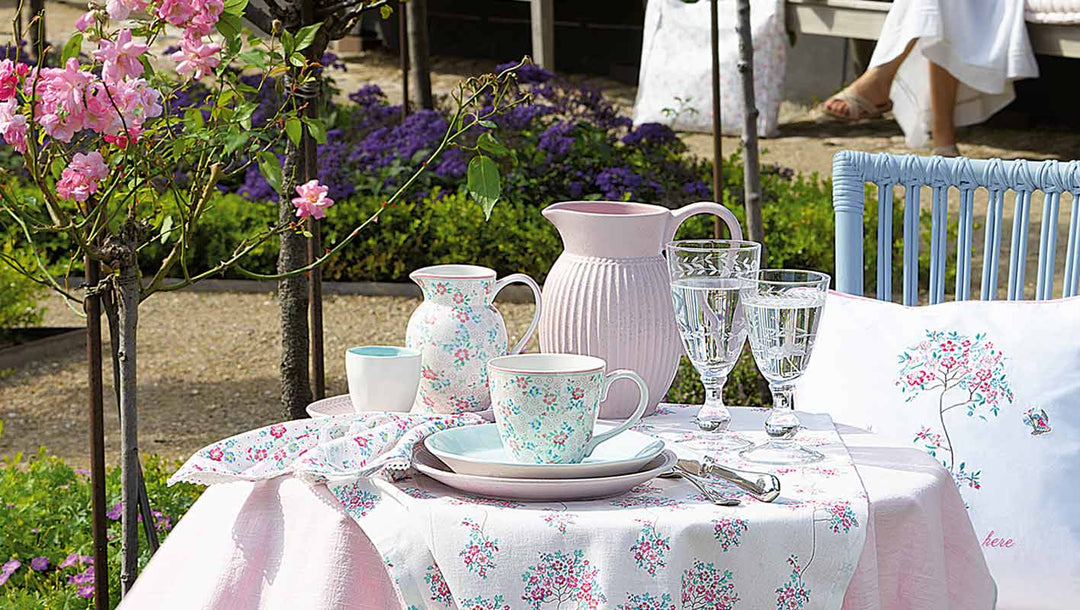 Ein Gartentisch mit Tellern, Tassen, einem Krug und Glaswaren mit Blumenmuster, umgeben von blühenden Blumen und viel Grün im Freien.