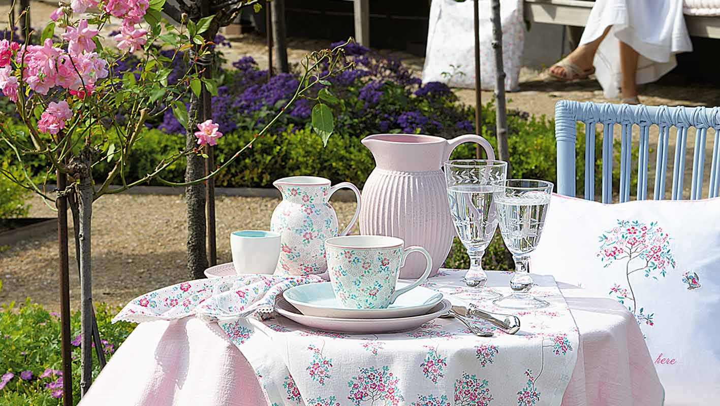 Ein Gartentisch mit Tellern, Tassen, einem Krug und Glaswaren mit Blumenmuster, umgeben von blühenden Blumen und viel Grün im Freien.