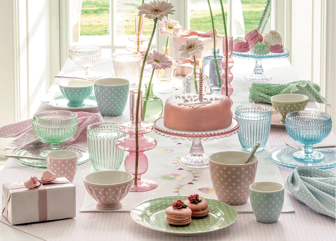 Ein pastellfarben gedeckter Tisch mit gepunktetem Geschirr, Glaswaren, einem rosa Kuchen mit Kerzen, Macarons, Blumen in Vasen und einem eingepackten Geschenk. Helles Fenster und Gartenblick im Hintergrund.
