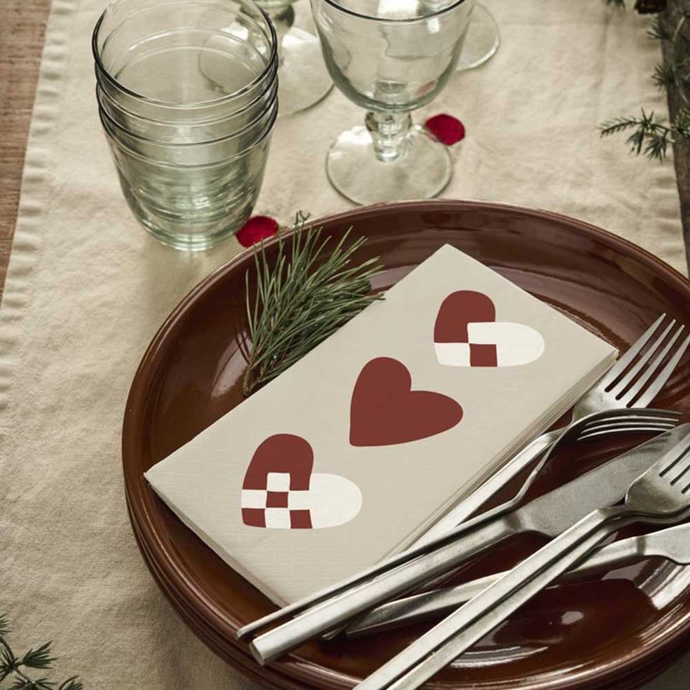 Ib-Laursen-Serviette mit roten Weihnachtsherzen auf einem braunen Teller mit Besteck und grünen Gläsern