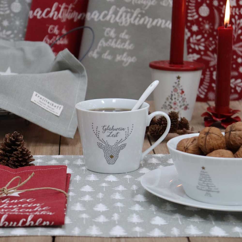 Glühweintasse mit Rentiermotiv auf einem weihnachtlich gedeckten Tisch mit roten Kerzen, Nüssen und Tannenzapfen