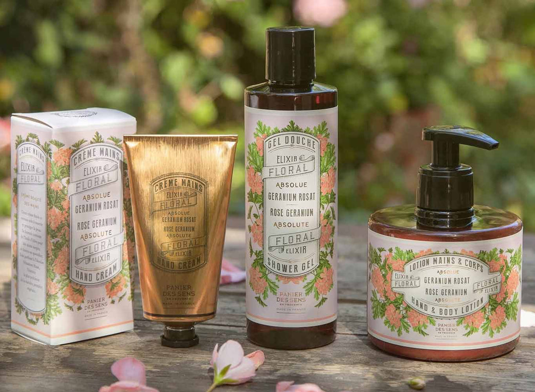 Vier florale Hautpflegeprodukte von Elixir Floral – Handcreme, Duschgel, Körperlotion und Verpackung – werden im Freien auf einer Holzoberfläche mit viel Grün im Hintergrund präsentiert.