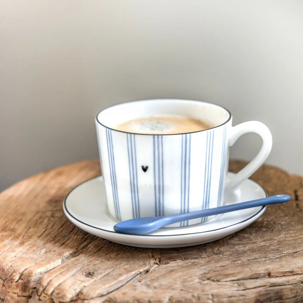 Eine gestreifte weiß-blaue Kaffeetasse mit Untertasse und blauem Löffel steht auf einer Holzfläche.