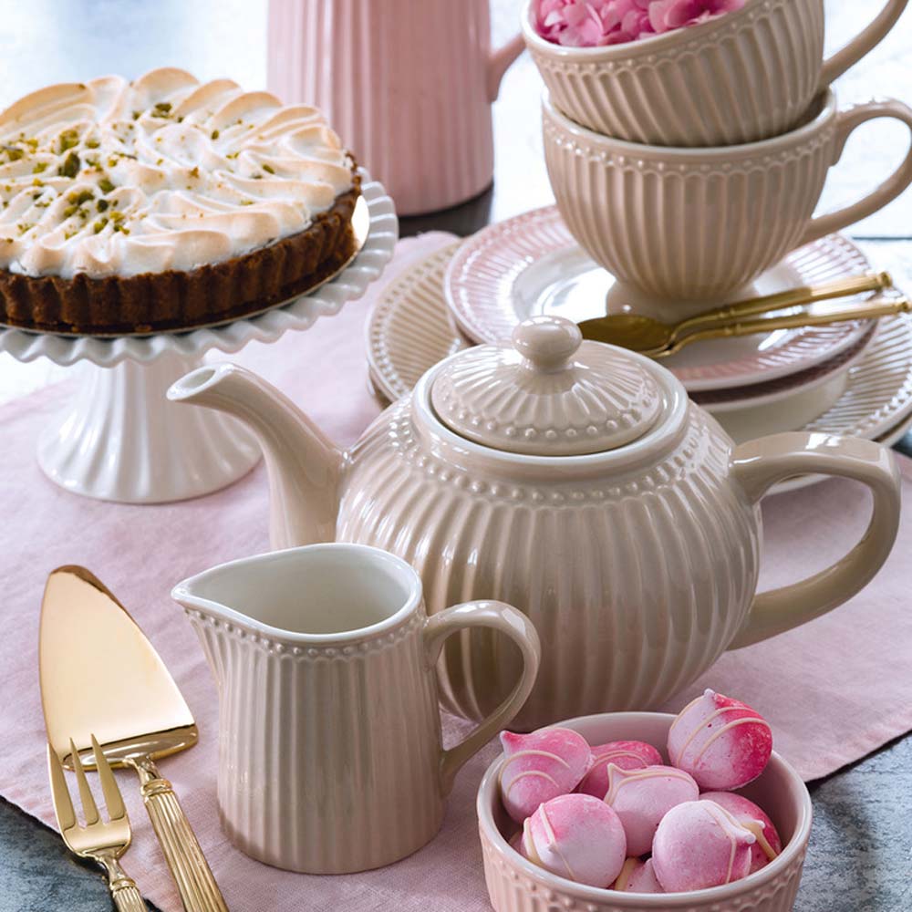 Farbenfrohes, pastellfarbenes Geschirr, darunter Tassen, Teller, Eierbecher, eine Teekanne und ein Krug, ordentlich auf einer weißen Oberfläche angeordnet, mit einer Schüssel Milch und roten Beeren, zwei Löffeln und einer Rose in einem Krug.