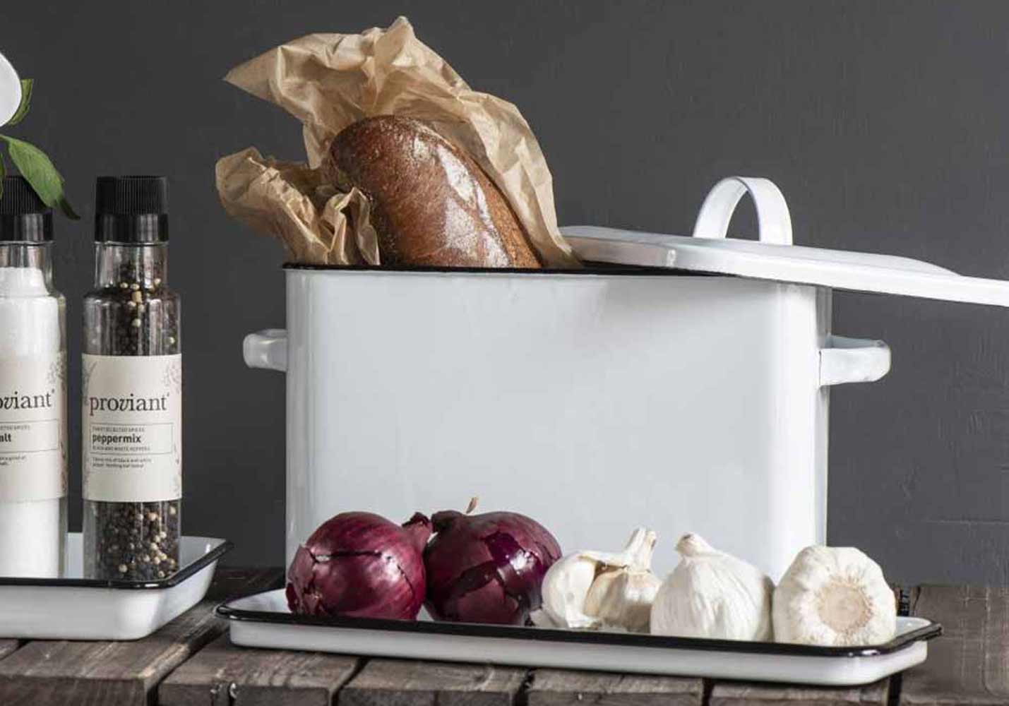 Ein weißer Emaille-Topf mit einem Laib Brot, umgeben von roten Zwiebeln, Knoblauch und Gewürzen auf einer Küchentheke.
