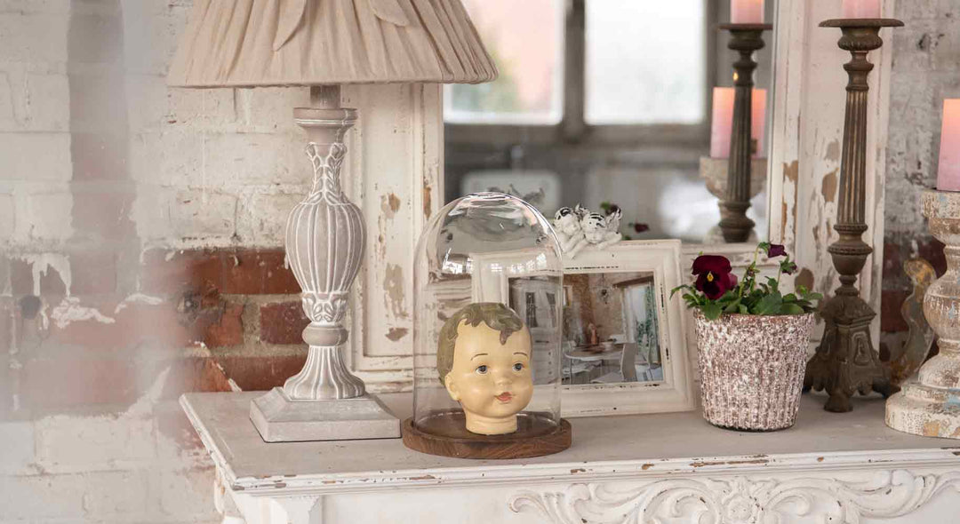 Der Kopf einer Babypuppe wird unter einer Glaskuppel auf einem weißen Kaminsims neben einer Lampe, gerahmten Fotos, Kerzen und einer Topfblume ausgestellt.