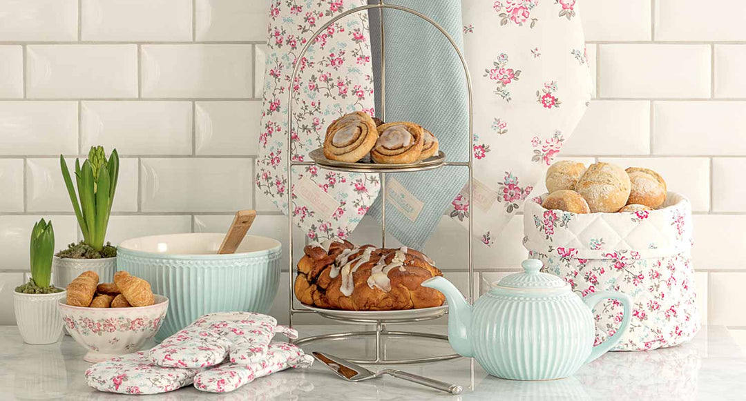 Ein mehrstöckiges Tablett mit Gebäck, einer Teekanne, einer Schüssel mit Schneebesen, Brötchen in einem gesteppten Korb, Topfhandschuhen und Topfpflanzen auf einer Marmorarbeitsplatte mit floralem Hintergrund.