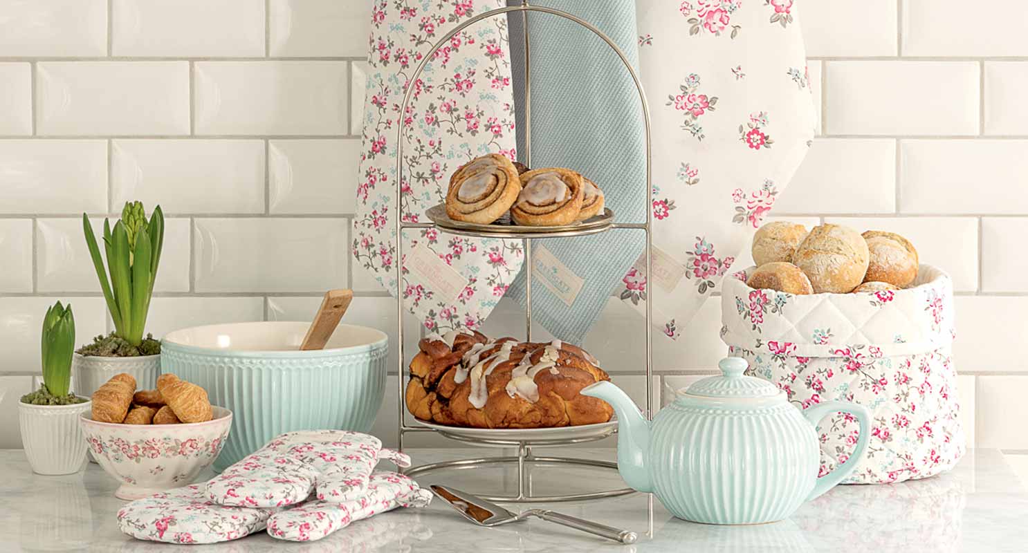 Ein mehrstöckiges Tablett mit Gebäck, einer Teekanne, einer Schüssel mit Schneebesen, Brötchen in einem gesteppten Korb, Topfhandschuhen und Topfpflanzen auf einer Marmorarbeitsplatte mit floralem Hintergrund.