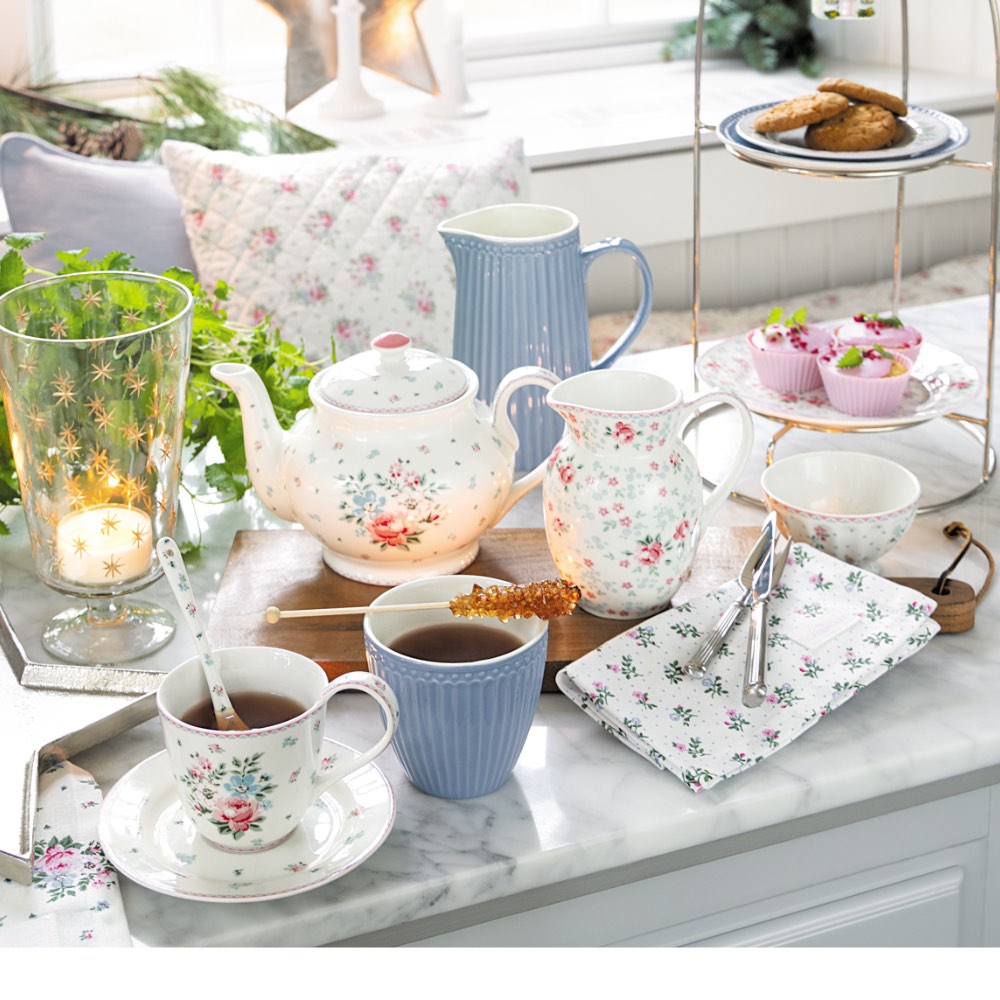 Auf einem Marmortisch in einem hellen Raum stehen ein Teeservice mit Blumenmuster, Tassen Tee, eine blaue Tasse, Kekse und Desserts, dazu Servietten, Pflanzen und Kerzen.
