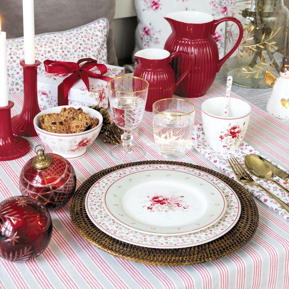 Ein gedeckter Tisch mit Geschirr mit Blumenmuster, Gläsern, goldenem Besteck, einem roten Krug, Kerzen, Dekoartikeln und einem verpackten Geschenk auf einer gestreiften Tischdecke.