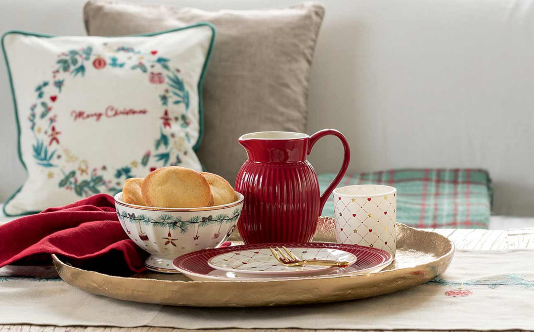 Auf einem goldenen Tablett stehen ein roter Krug, eine Schüssel mit Keksen, ein Teller mit einer Gabel, eine Tasse und eine rote Serviette. Dahinter ist ein festliches Kissen mit der Aufschrift „Frohe Weihnachten“ zu sehen.