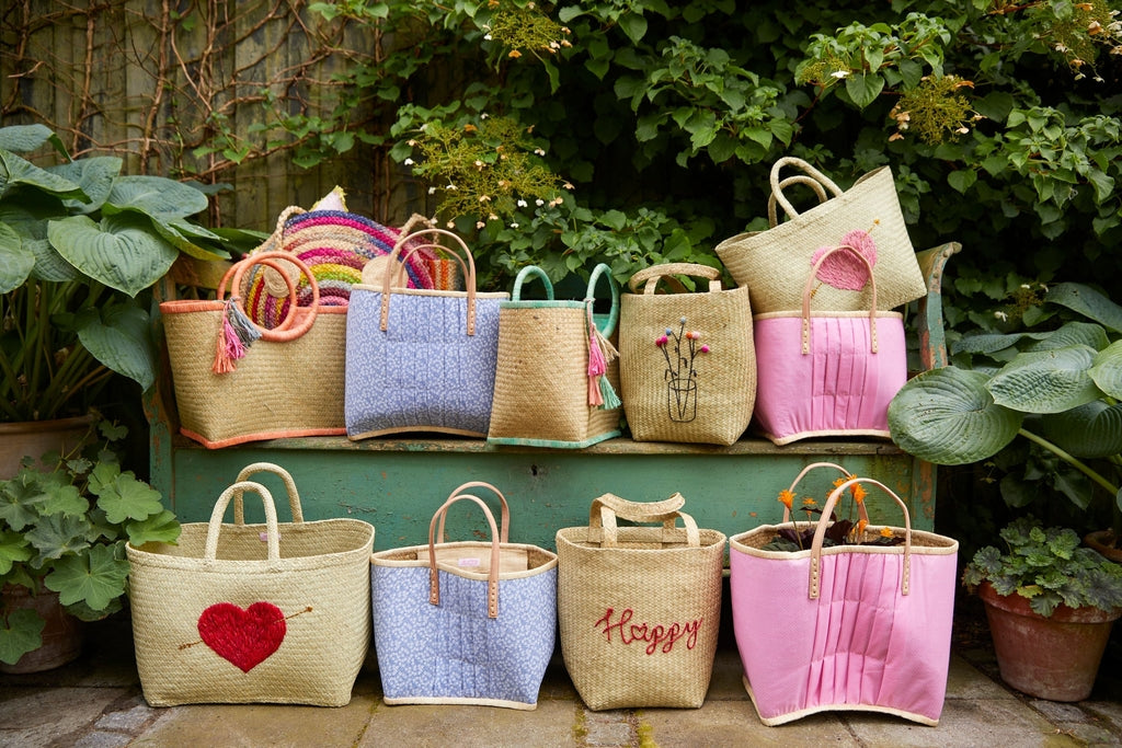 Eine Sammlung bunter gewebter Strohtaschen mit verschiedenen Designs und Mustern wird im Freien auf und vor einer grünen Bank, umgeben von Blattpflanzen, ausgestellt.
