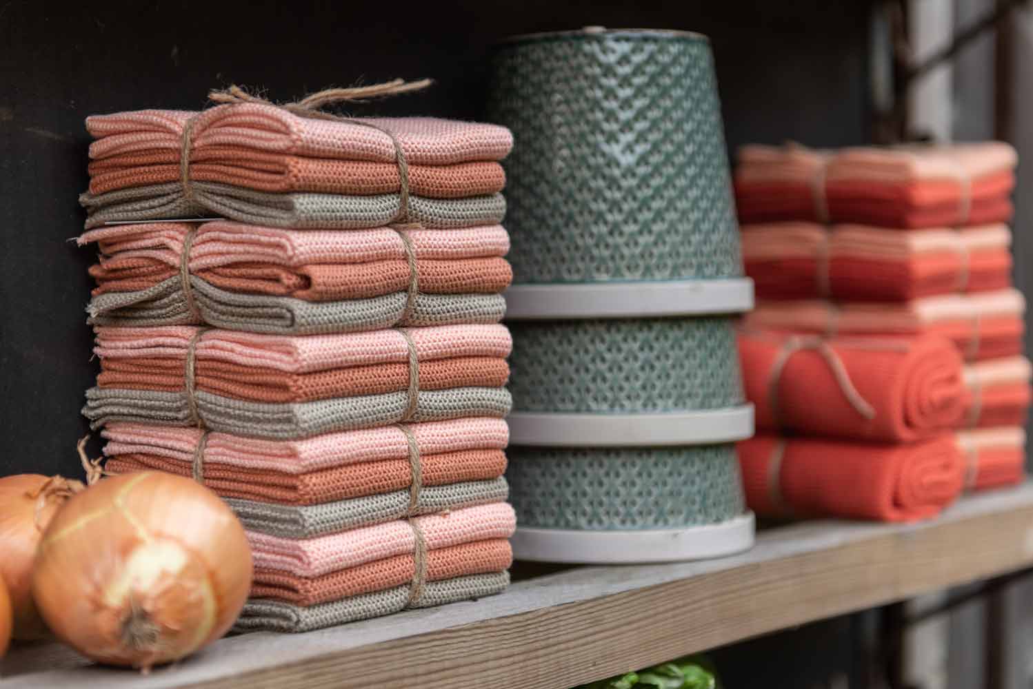 Stapel gefalteter, zusammengebundener Geschirrtücher in Rosa und Beige, drei strukturierte grüne Blumentöpfe und ein paar Zwiebeln, angeordnet auf einem Holzregal.