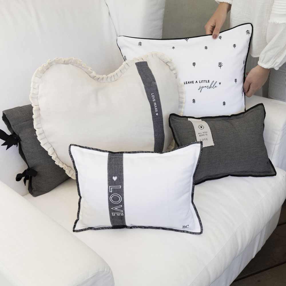 Eine Kollektion von **Bastion Collection - Kissen Love 25 x 35 cm mit Füllung** auf einem weißen Sofa. Die Kissen haben verschiedene Farben und Designs, darunter Herzformen und Streifenmuster. Rechts ist teilweise eine Person zu sehen.
