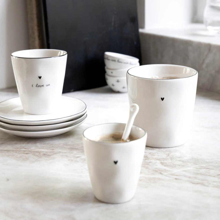 Die Becher und Teller von Bastion Collections in natürlicher Form sind auf einer Marmorplatte angeordnet. Die Tassen sind mit kleinen Herzen und Sprüchen wie „Ich liebe uns“ versehen. Im Vordergrund liegt ein Löffel in einer Tasse mit Getränk.