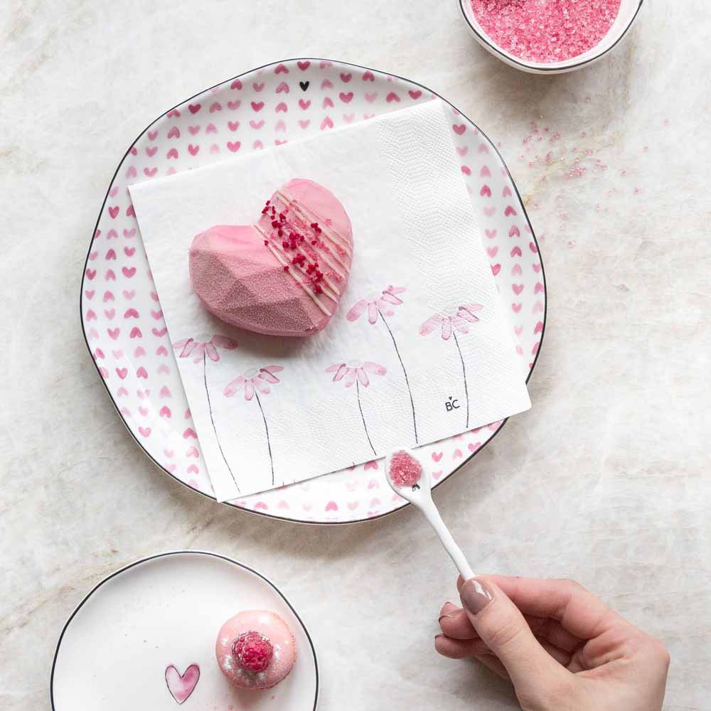 Eine Hand hält einen Löffel mit rosa Zucker über einem angebissenen herzförmigen rosa Dessert auf einem Bastion Collections - Frühstücksteller Rose Hearts all over, auf einer Serviette mit Blumenmuster. Rosa Zucker und ein rundes Dessert stehen daneben.