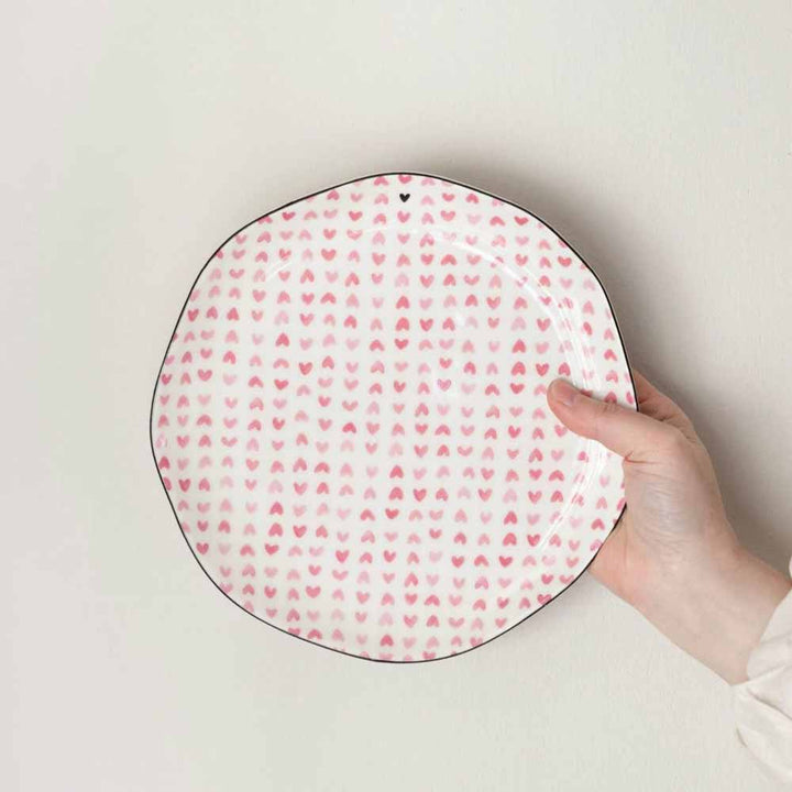 Eine Hand hält den Bastion Collections Frühstücksteller Rose Hearts all over, einen weißen Keramikteller mit kleinen rosa Herzmustern und einem leicht unregelmäßigen, schwarz umrandeten Rand.