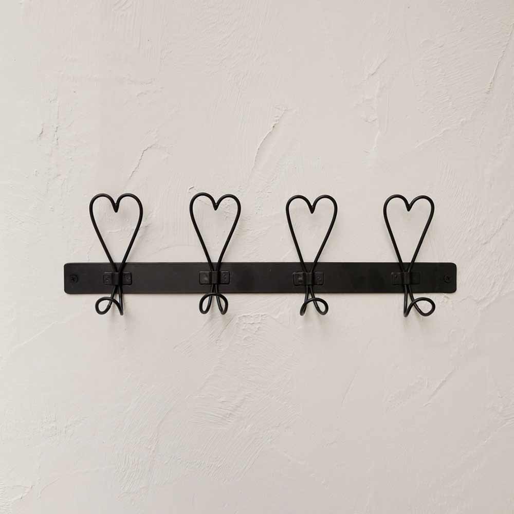 Die Garderobe der Bastion Collections mit vier herzförmigen Drahthaken, die an einer glatten schwarzen Metallstange befestigt sind, ist elegant an einer strukturierten weißen Wand montiert.