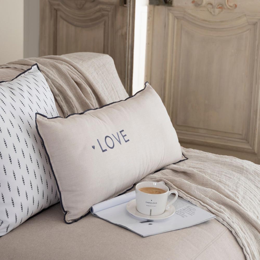 Ein beiges Kissen mit der Aufschrift „LOVE“ (Bastion Collections – Kissenbezug Chambray Hearts 50 x 50 cm) liegt auf einem Sofa mit einer Decke. Dahinter befindet sich ein weißes Kissen. In der Nähe steht eine Kaffeetasse auf einer aufgeschlagenen Zeitschrift, eingerahmt von verzierten Holztüren im Hintergrund.