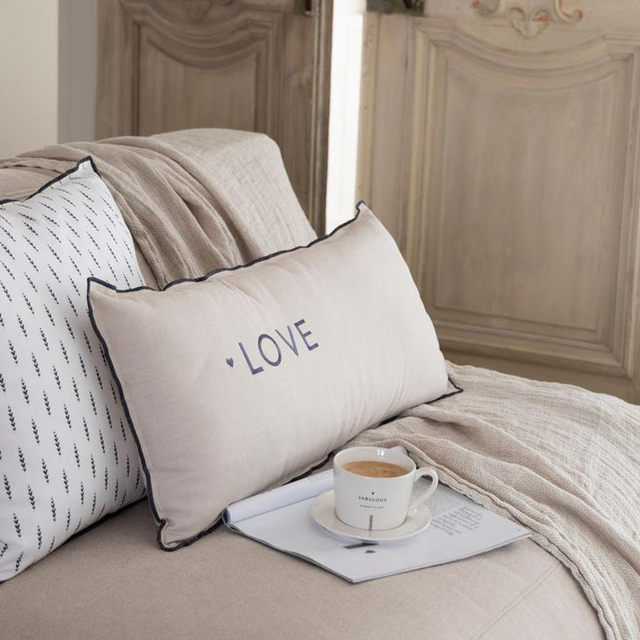 Ein beiges Kissen mit der Aufschrift „LOVE“ (Bastion Collections – Kissenbezug Chambray Hearts 50 x 50 cm) liegt auf einem Sofa mit einer Decke. Dahinter befindet sich ein weißes Kissen. In der Nähe steht eine Kaffeetasse auf einer aufgeschlagenen Zeitschrift, eingerahmt von verzierten Holztüren im Hintergrund.