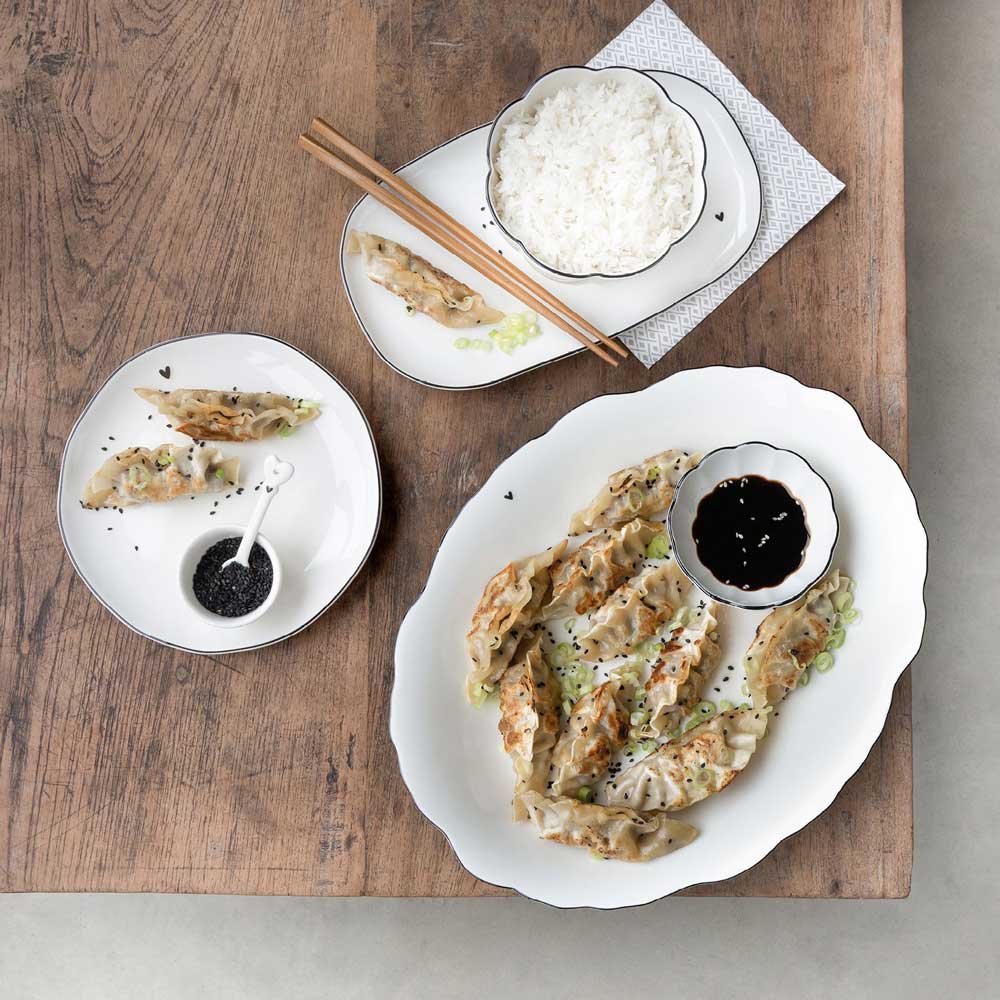 Teigtaschen auf einer Servierplatte „White Flower“ von Bastion Collections, garniert mit Sojasauce und Sesamkörnern, neben einer Schüssel Reis auf einem Holztisch.