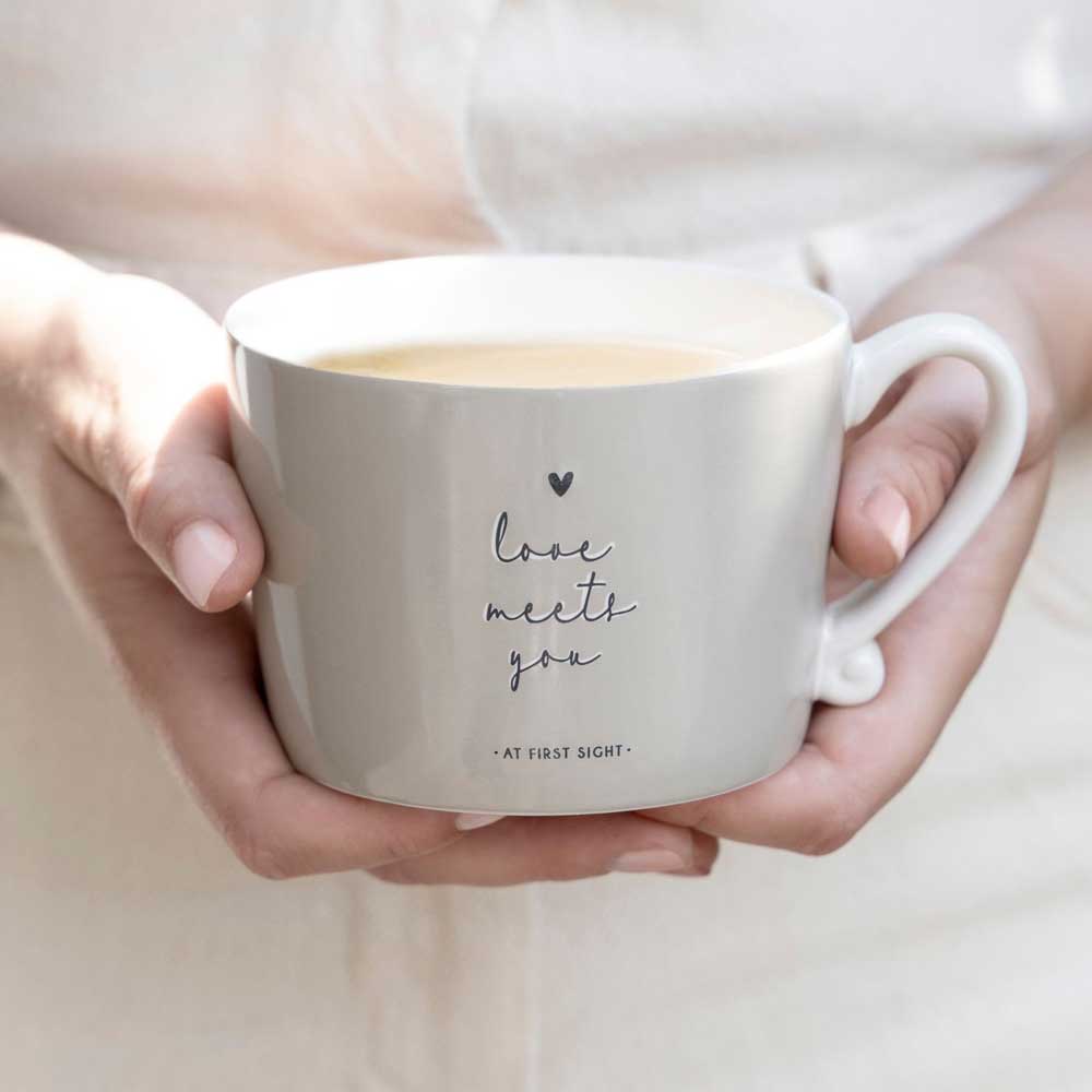 Eine Person hält die Bastion Collections - Tasse Love meets you Titane, eine weiße Tasse, die mit einem leichten Getränk gefüllt ist. Auf der Tasse steht "Liebe begegnet dir auf den ersten Blick" und ein kleines Herz über dem Text.
