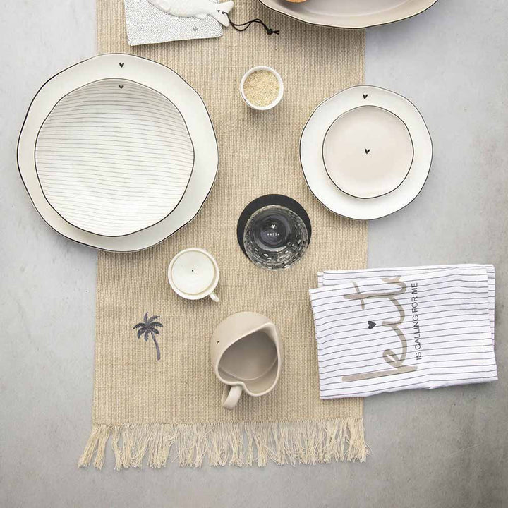 Ein Set aus weißem Geschirr, einer Tasse, kleinen Schüsseln und einer schwarzen Kerze auf einem Tischläufer aus Leinen „Palm Tree“ von Bastion Collections. Daneben liegt ein Tischtuch mit schwarzen Streifen und teilweisem Text.