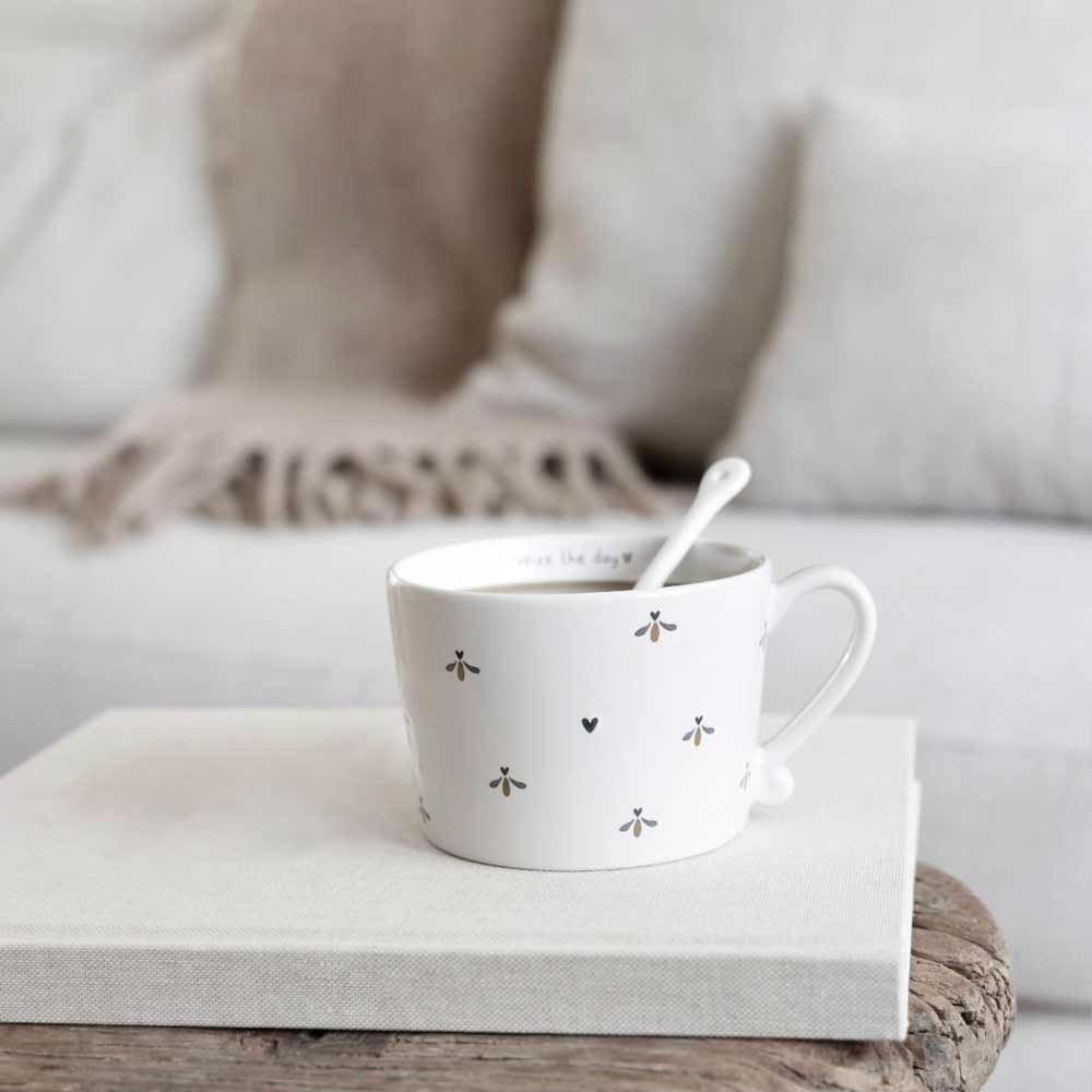 Ein wellenförmiger weißer Becher von Bastion Collections – Löffel für Kaffee mit einem kleinen Löffel darin steht auf einer gefalteten weißen Decke auf einer Holzoberfläche, im Hintergrund liegt eine beigefarbene Decke auf einem hellgrauen Sofa.