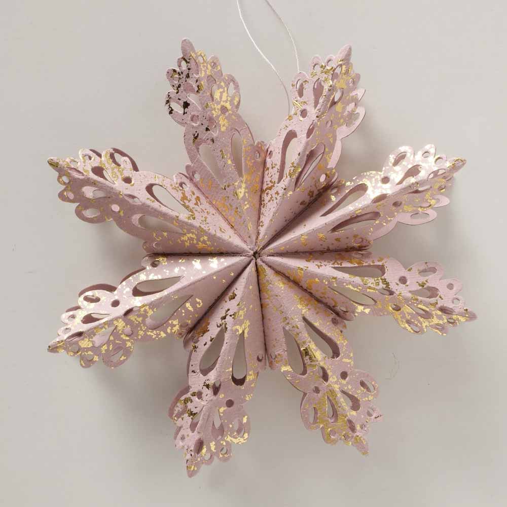 Eine hängende 3D-Schneeflockendekoration von Boltze mit komplizierten ausgeschnittenen Mustern und goldenen Sprenkeln auf einem hellrosa Hintergrund.