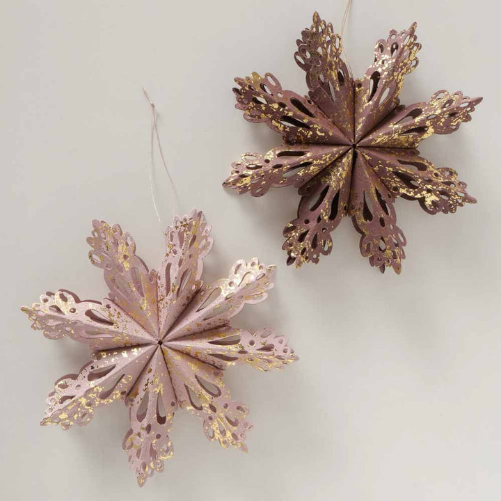 Zwei 3D-Schneeflocken von Boltze mit komplizierten ausgeschnittenen Mustern in Rosa- und Brauntönen mit goldenen Sprenkeln, hängen vor einem hellen Hintergrund.