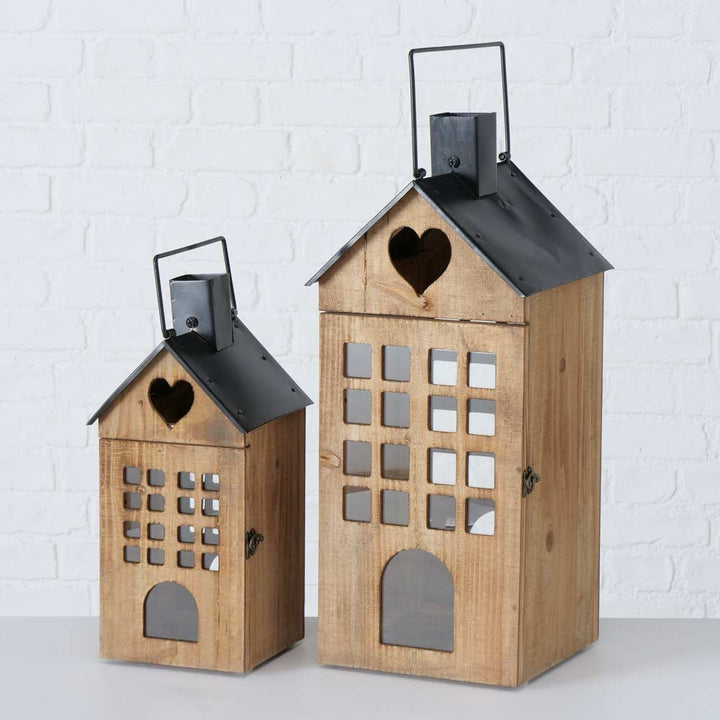 Zwei Holzlaternen „Boltze – Laterne Brixen Haus“ mit schwarzen Metalldächern haben die Form von Häusern, jeweils mit herzförmigen Ausschnitten, quadratischen Fenstern und Türausschnitten vor einer weißen Backsteinmauer.