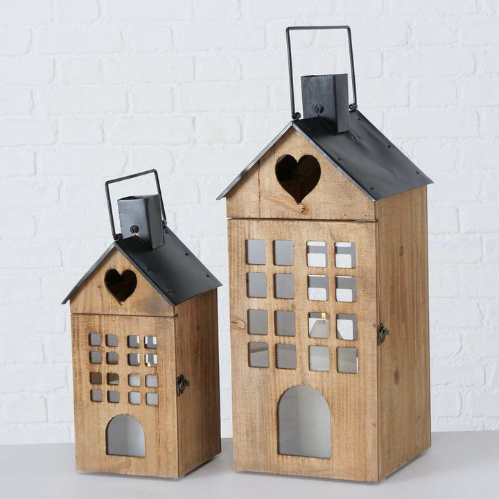 Zwei hölzerne Laternen von Boltze - Laterne Brixen Haus in Form von Häusern haben schwarze Metalldächer. Die größere Laterne hat einen herzförmigen Ausschnitt und mehrere quadratische Fenster. Beide sind vor einer weißen Backsteinmauer ausgestellt.