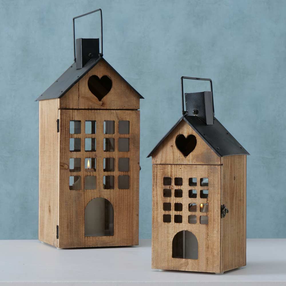 Zwei Boltze - Laterne Brixen Haus Holzlaternen in Form kleiner Häuser mit schwarzen Metalldächern und herzförmigen Ausschnitten oben, vor einem hellblauen Hintergrund. Die Laternen haben Gitter und gewölbte Fenster.