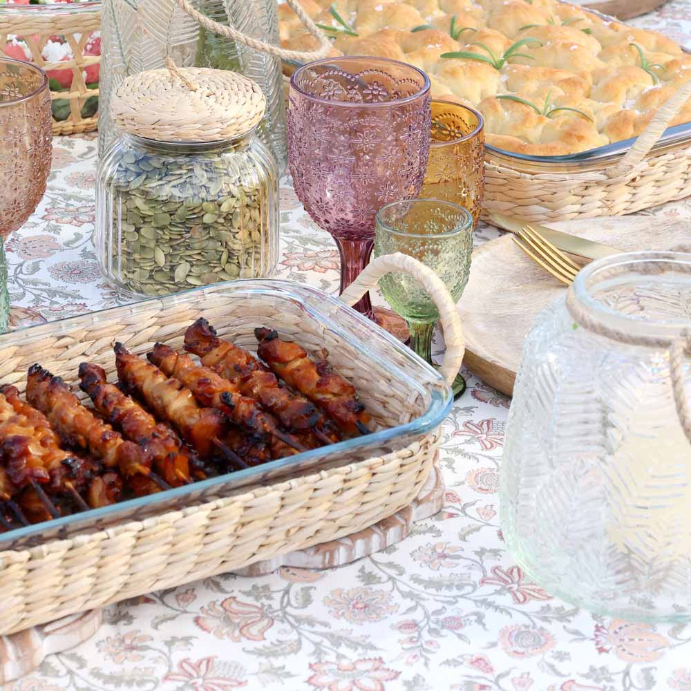 Die Chic Antique - Auflaufform mit abnehmbarem Flechtkorb enthält aufgespießtes Grillfleisch, während bunte Gläser, ein Glas mit Samen und ein Tablett mit Focaccia auf einer gemusterten Tischdecke stehen.
