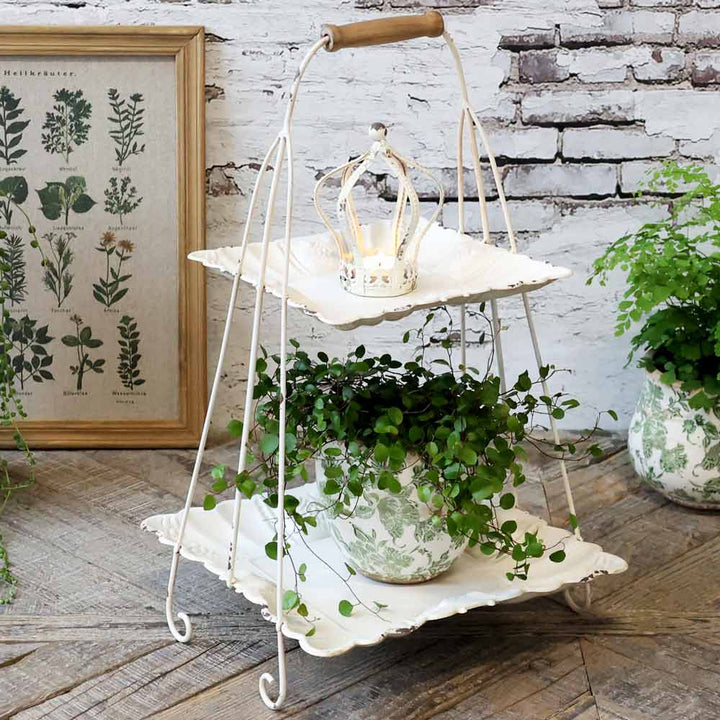 Zweistöckiger weißer Metallständer mit einer kleinen Krone als Ornament oben drauf. Auf der unteren Etage steht eine Topfpflanze mit grünem Blattwerk. Im Hintergrund sind ein gerahmter botanischer Druck und eine weitere Pflanze zu sehen. Chic Antique - Etagere mit Henkel