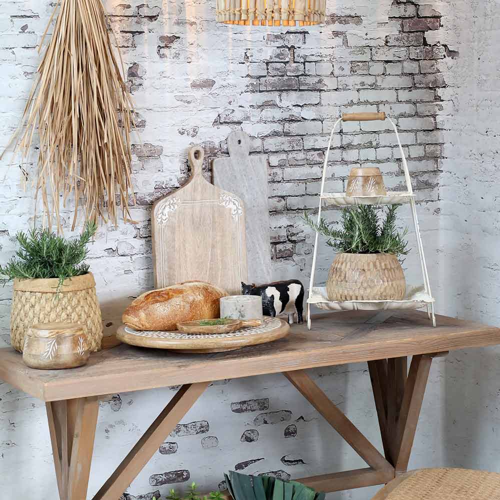 Ein rustikaler Tisch vor einer weißen Backsteinwand, präsentiert mit Schneidebrettern, Brot, einer Kuhfigur, Topfpflanzen und einer Chic Antique – Etagere mit Henkel mit zusätzlichem Dekor.