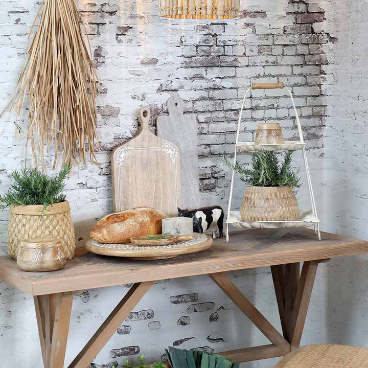 Ein rustikaler Tisch vor einer weißen Backsteinwand, präsentiert mit Schneidebrettern, Brot, einer Kuhfigur, Topfpflanzen und einer Chic Antique – Etagere mit Henkel mit zusätzlichem Dekor.