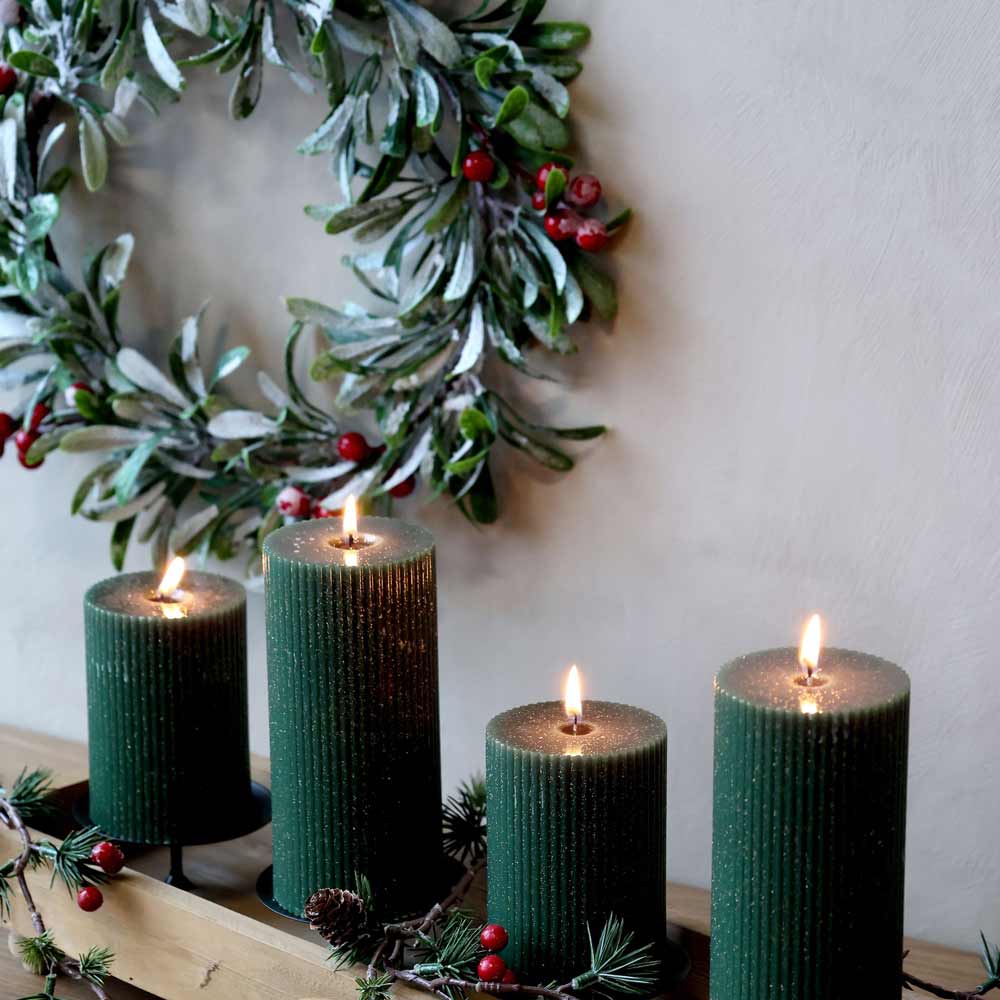 Vier brennende grüne Stumpenkerzen auf einem Holzregal mit dem Chic Antique Fleur-Kranz, verziert mit Schnee und roten Beeren im Hintergrund.