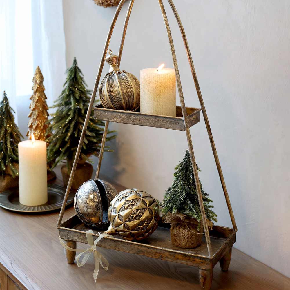Ein Chic Antique Fleur Tannenbaum mit Glitzergold, geschmückt mit festlichen Ornamenten und Kerzen, platziert auf einem rustikalen Metallregal neben kleinen künstlichen Weihnachtsbäumen auf einer Holzoberfläche.