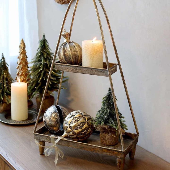Ein Chic Antique Fleur Tannenbaum mit Glitzergold, geschmückt mit festlichen Ornamenten und Kerzen, platziert auf einem rustikalen Metallregal neben kleinen künstlichen Weihnachtsbäumen auf einer Holzoberfläche.