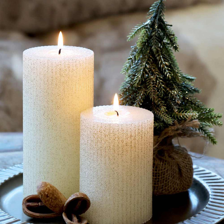 Neben dem Chic Antique – Fleur Tannenbaum mit Glitzergold stehen zwei brennende weiße Kerzen auf einer metallischen Oberfläche.