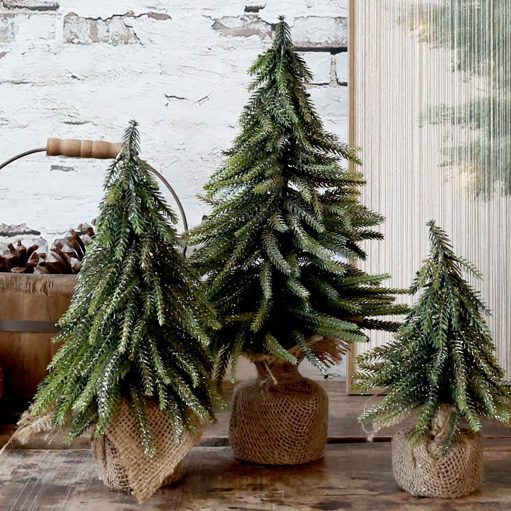 Drei Chic Antique - Fleur Tannenbaum mit Glitzergold-Bäumen sind elegant in Leinensäcke verpackt und auf einer Holzfläche vor einer weißen Ziegelwand ausgestellt.