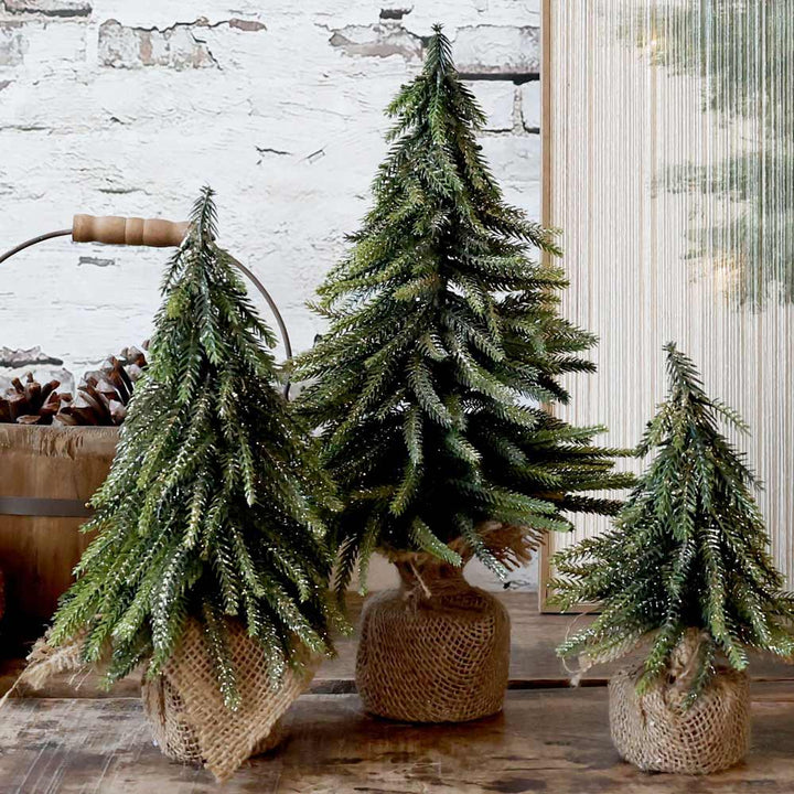 Drei Chic Antique - Fleur Tannenbaum mit Glitzergold-Bäumen sind elegant in Leinensäcke verpackt und auf einer Holzfläche vor einer weißen Ziegelwand ausgestellt.