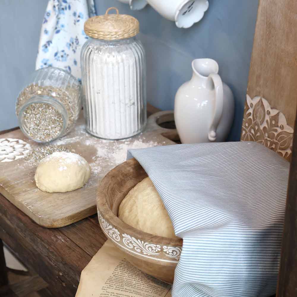 Küchenablage mit hohem gerilltem Vorratsglas, hellem Krug und einer Holzschale mit Teig unter einem fein blau gestreiften Tuch